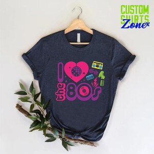 May include: A dark blue t-shirt with a graphic design featuring a pink heart with a disco ball inside, the text "I love the 80s" in purple and pink, and 80s icons like roller skates, sunglasses, cassette tapes, and a record.