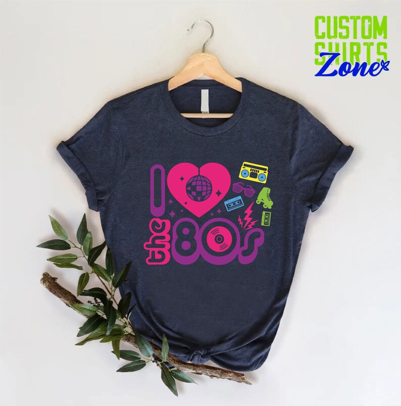 May include: A dark blue t-shirt with a graphic design featuring a pink heart with a disco ball inside, the text "I love the 80s" in purple and pink, and 80s icons like roller skates, sunglasses, cassette tapes, and a record.