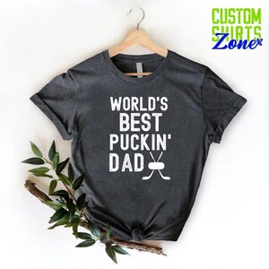May include: A dark gray t-shirt with the text "World's Best Puckin' Dad" in white with a hockey stick and puck graphic.
