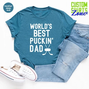May include: A teal blue t-shirt with the text "WORLD'S BEST PUCKIN' DAD" in white with a hockey stick and puck graphic.