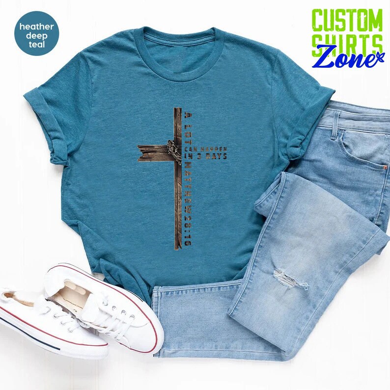 May include: A teal blue t-shirt with a wood-grain cross design. The cross has a crown of thorns and the text "ALL THINGS CAN HAPPEN IN 3 DAYS" and "MATTHEW 28:16".