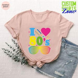 May include: A pink t-shirt with the text "I love the 80's" in rainbow colors. The design includes a heart, a cassette tape, and sunglasses.
