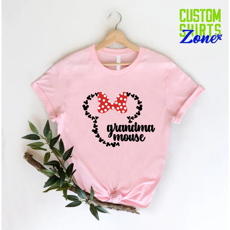Op de afbeelding: Een lichtroze t-shirt met een Minnie Mouse-ontwerp. Het ontwerp bevat een rode strik met witte stippen, zwarte muizenoren en de woorden "grandma mouse" in zwart schrift. Het t-shirt hangt aan een houten hanger.