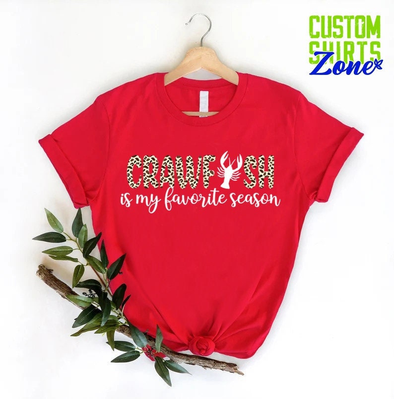 Puede incluir: Camiseta roja con un gr&aacute;fico blanco que dice "Crawfish is my favorite season". El gr&aacute;fico est&aacute; en un patr&oacute;n de estampado de leopardo e incluye una ilustraci&oacute;n de cangrejo de r&iacute;o blanco.