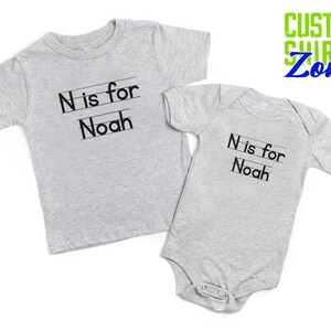 May include: Two gray t-shirts with the text "N is for Noah" printed on them. The shirts are in different sizes, one is a long-sleeved shirt and the other is a short-sleeved bodysuit.