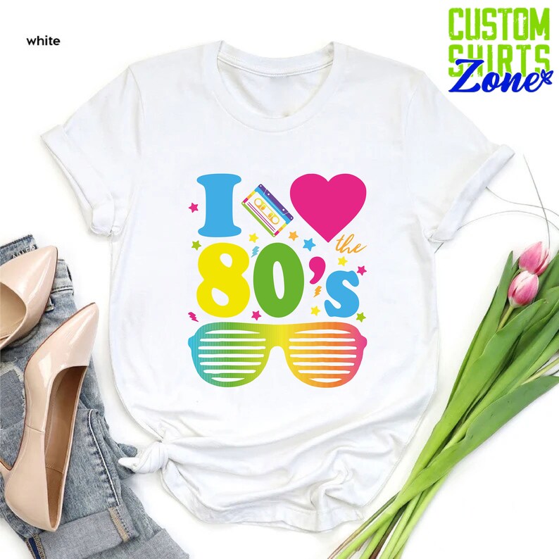 May include: White t-shirt with a graphic design that says "I love the 80's" with a cassette tape, heart, and sunglasses.