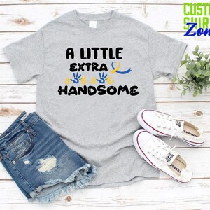 Pode incluir: Uma camiseta cinza com o texto "A Little Extra Handsome" em preto. O texto &eacute; cercado por tr&ecirc;s impress&otilde;es de m&atilde;os amarelas e uma fita azul.
