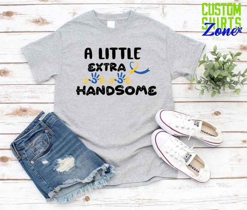 Pode incluir: Uma camiseta cinza com o texto "A Little Extra Handsome" em preto. O texto &eacute; cercado por tr&ecirc;s impress&otilde;es de m&atilde;os amarelas e uma fita azul.