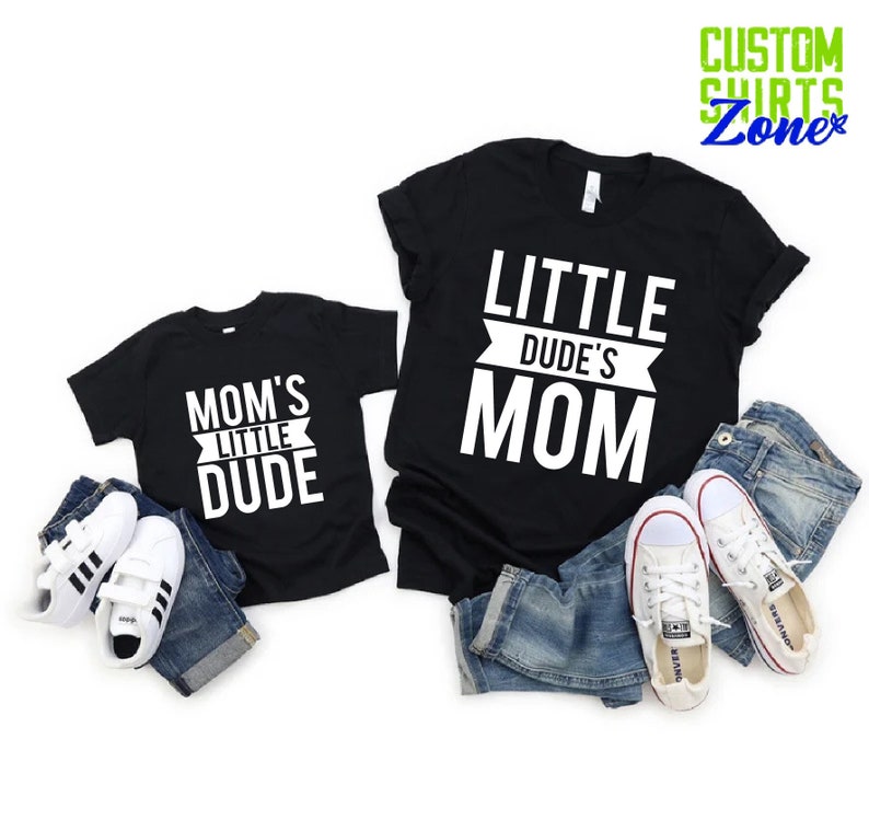 Op de afbeelding: Twee zwarte T-shirts met witte tekst: "MOM'S LITTLE DUDE" en "LITTLE DUDE'S MOM". De shirts worden getoond met denim shorts en witte sneakers. De afbeelding bevat ook de tekst "CUSTOM SHIRTS Zone" in groen en blauw.