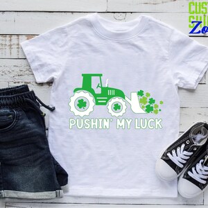May include: White t-shirt with a green tractor pushing a pile of green shamrocks. The text "Pushin' My Luck" is printed below the tractor.