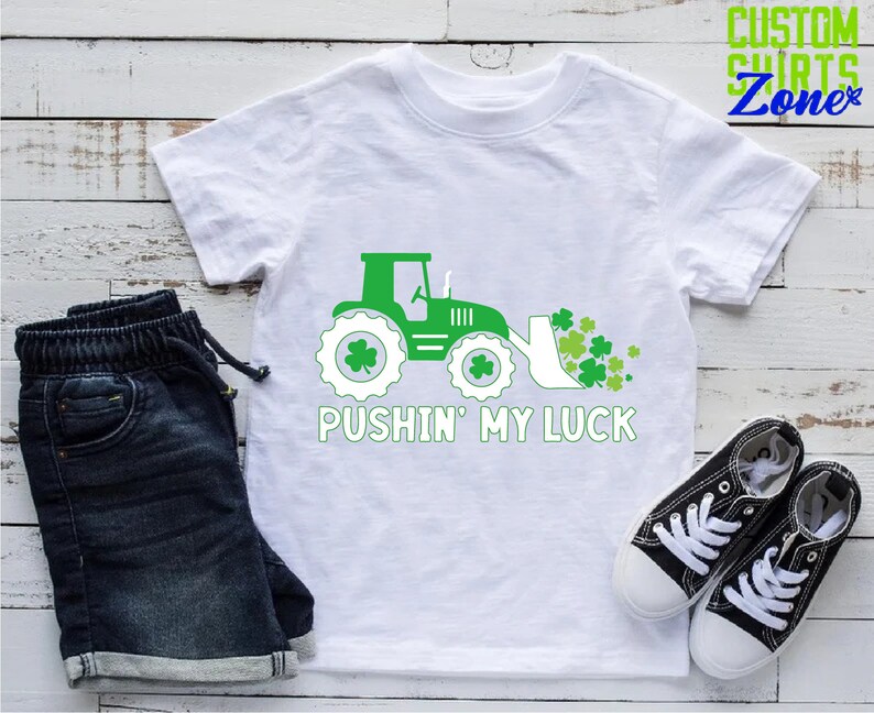 May include: White t-shirt with a green tractor pushing a pile of green shamrocks. The text "Pushin' My Luck" is printed below the tractor.