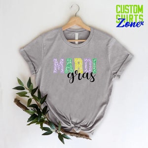 May include: A gray t-shirt with the words "Mardi Gras" printed on it in purple, green, and yellow letters with a leopard print pattern.