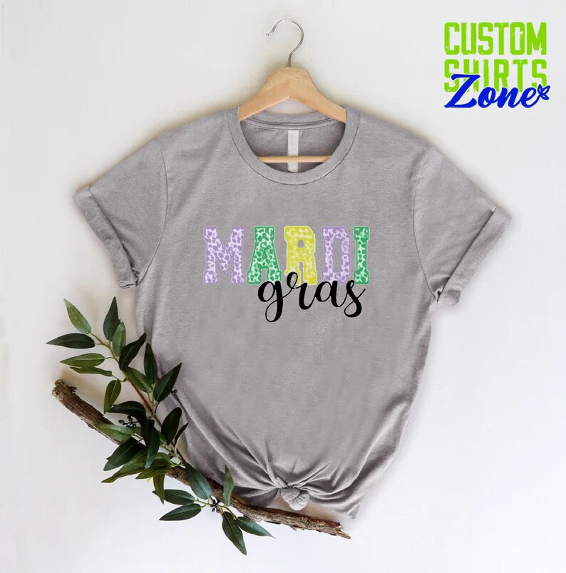 May include: A gray t-shirt with the words "Mardi Gras" printed on it in purple, green, and yellow letters with a leopard print pattern.