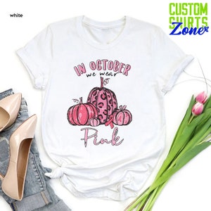 May include: White t-shirt with a pink and black graphic of pumpkins and a pink ribbon. The text "In October we wear Pink" is printed on the shirt.