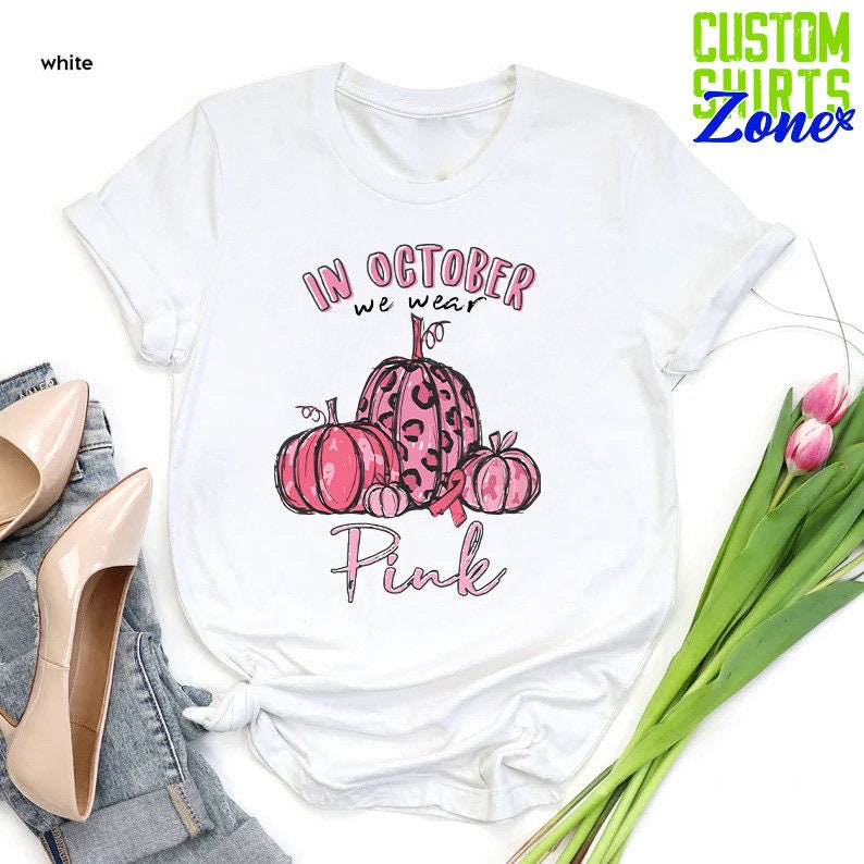 May include: White t-shirt with a pink and black graphic of pumpkins and a pink ribbon. The text "In October we wear Pink" is printed on the shirt.