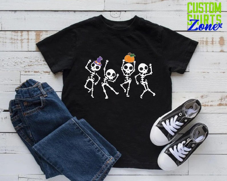 May include: Black t-shirt with a graphic of four skeletons dancing. The skeletons are wearing hats and holding pumpkins. The shirt is on a white wooden surface with a pair of blue jeans and white sneakers.