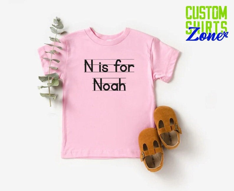 May include: Pink short-sleeved t-shirt with the text "N is for Noah" printed on the front.