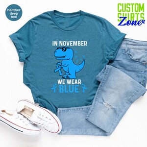 May include: Teal t-shirt with a blue dinosaur wearing sunglasses and a blue ribbon. The text reads "IN NOVEMBER WE WEAR BLUE." The shirt is paired with light blue jeans and white sneakers. The shirt is part of the Custom Shirts Zone collection.