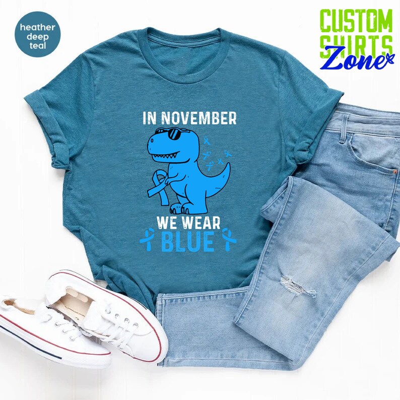 May include: Teal t-shirt with a blue dinosaur wearing sunglasses and a blue ribbon. The text reads "IN NOVEMBER WE WEAR BLUE." The shirt is paired with light blue jeans and white sneakers. The shirt is part of the Custom Shirts Zone collection.