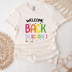 以下が含まれることがあります： 「WELCOME BACK TO SCHOOL!」のカラフルな文字がプリントされたクリーム色のTシャツ。テキストには星と「Mrs. Davis」の名前が下部に含まれています。柔らかい素材で作られたシャツです。