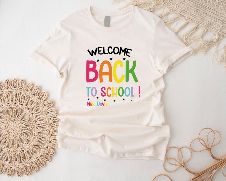 以下が含まれることがあります： 「WELCOME BACK TO SCHOOL!」のカラフルな文字がプリントされたクリーム色のTシャツ。テキストには星と「Mrs. Davis」の名前が下部に含まれています。柔らかい素材で作られたシャツです。