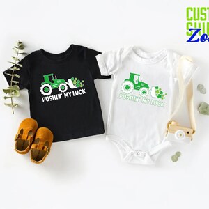May include: Two black and white shirts with a green tractor and shamrocks. The shirts say "Pushin' My Luck".