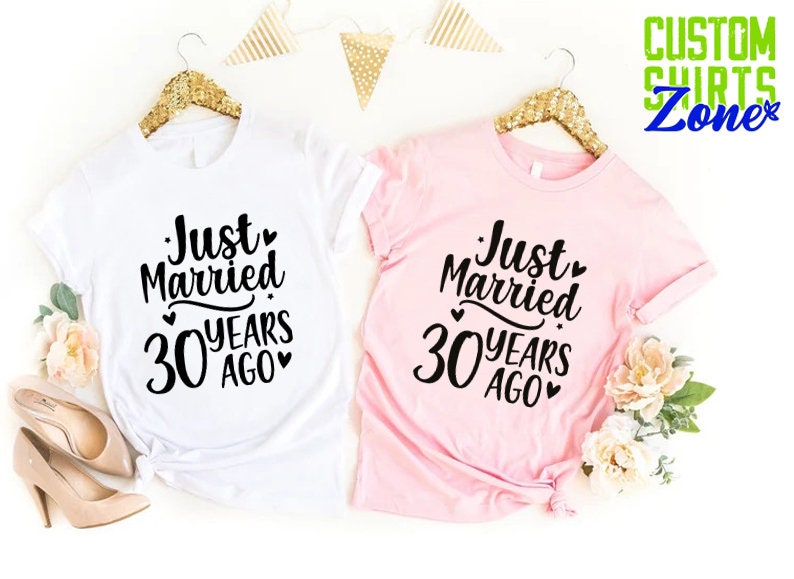 May include: Two t-shirts, one white and one pink, each with the text "Just Married 30 Years Ago" in black script. The shirts are displayed on gold hangers, with floral and decorative elements.