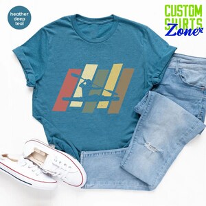 K&ouml;nnte beinhalten: Ein blaues T-Shirt mit einem Vintage-Grafikdruck einer Person im Kajak. Die Grafik besteht aus f&uuml;nf vertikalen Streifen in den Farben Rot, Orange, Gelb, Gr&uuml;n und Blau. Der Text "CUSTOM SHIRTS Zone" befindet sich in der oberen rechten Ecke des Bildes.