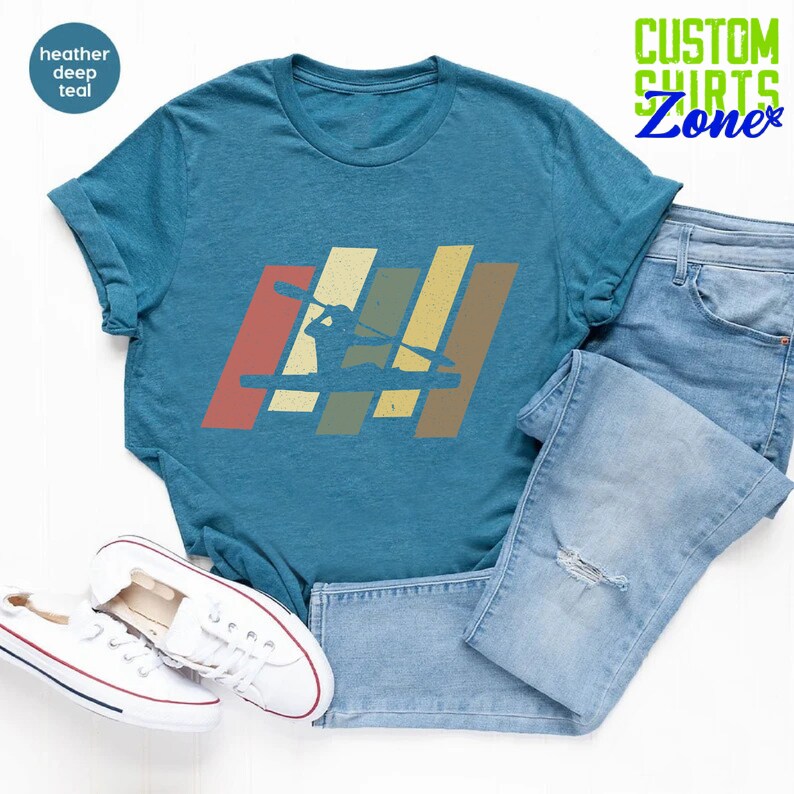 K&ouml;nnte beinhalten: Ein blaues T-Shirt mit einem Vintage-Grafikdruck einer Person im Kajak. Die Grafik besteht aus f&uuml;nf vertikalen Streifen in den Farben Rot, Orange, Gelb, Gr&uuml;n und Blau. Der Text "CUSTOM SHIRTS Zone" befindet sich in der oberen rechten Ecke des Bildes.