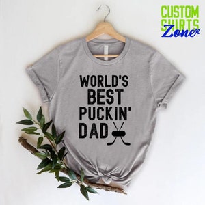 May include: A gray t-shirt with the text "WORLD'S BEST PUCKIN' DAD" and a hockey stick and puck graphic.