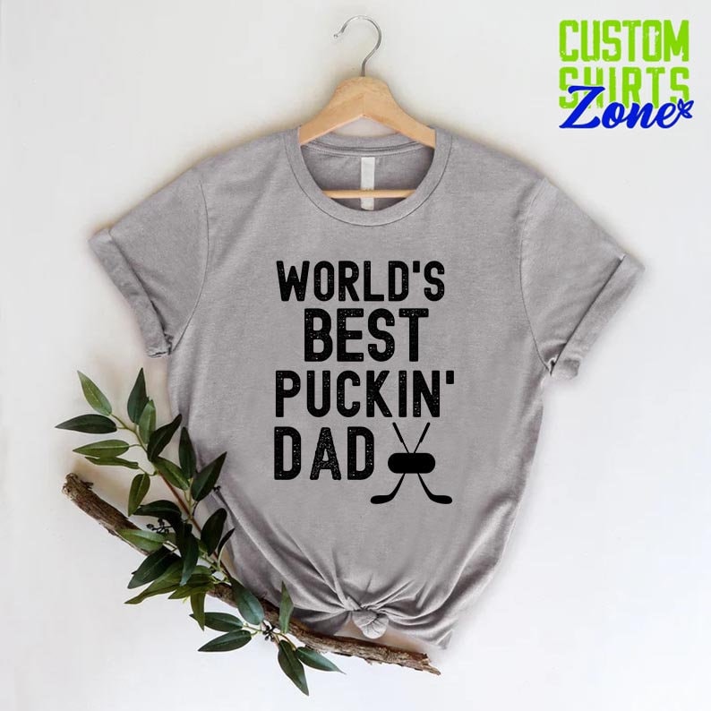 May include: A gray t-shirt with the text "WORLD'S BEST PUCKIN' DAD" and a hockey stick and puck graphic.