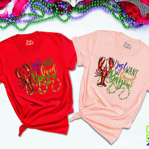 May include: Two t-shirts with a Mardi Gras theme. The red shirt says "I just want to be loved in the Bayou" and the pink shirt says "I just want to be loved in the Bayou". Both shirts feature a crawfish and beads.