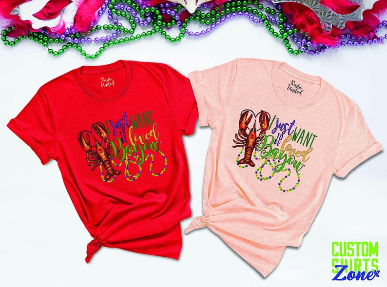 May include: Two t-shirts with a Mardi Gras theme. The red shirt says "I just want to be loved in the Bayou" and the pink shirt says "I just want to be loved in the Bayou". Both shirts feature a crawfish and beads.