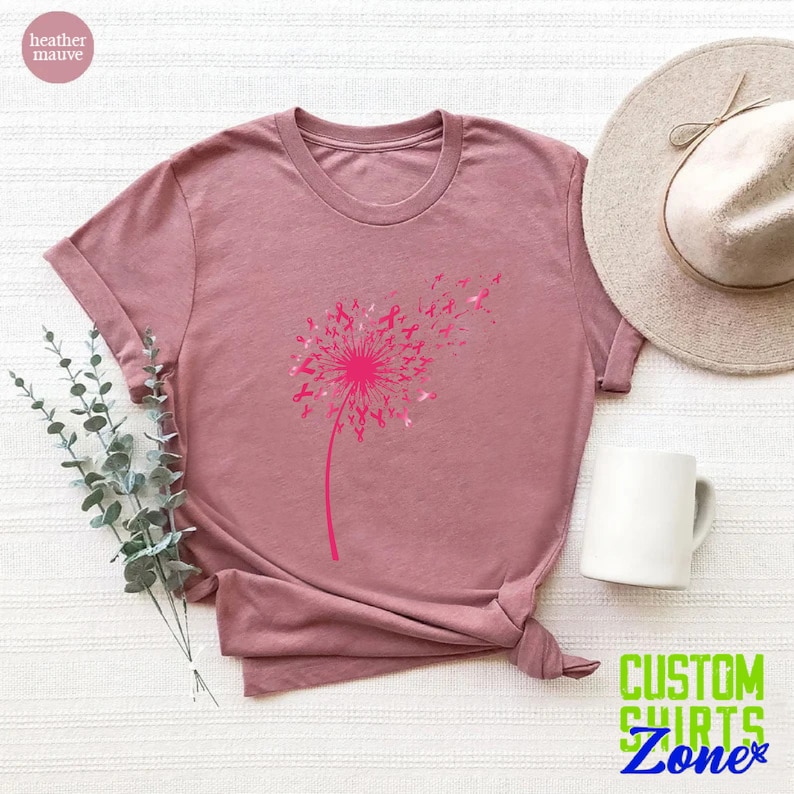 Op de afbeelding: Een heide-paarse t-shirt met een roze paardenbloemontwerp. De paardenbloem is gemaakt van roze linten, ter symbolisering van borstkankerbewustzijn.