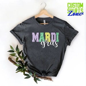 May include: A dark gray t-shirt with the words "Mardi Gras" printed on the front. The letters are in a purple, green, and yellow color scheme with a leopard print pattern.