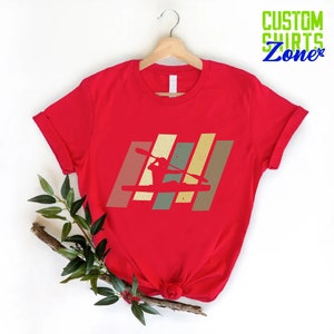 K&ouml;nnte beinhalten: Rotes T-Shirt mit einer Retro-Grafik einer Person im Kajak. Die Grafik besteht aus vertikalen Streifen in Braun-, Gr&uuml;n- und Gelbt&ouml;nen.