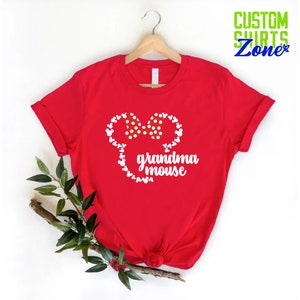 Op de afbeelding: Een rood t-shirt met een witte muizenkop met een stippen strik en de woorden "oma muis". Het shirt hangt aan een houten hanger, met een takje groen op de voorgrond. De tekst "CUSTOM SHIRTS Zone" staat rechtsboven.