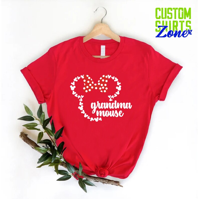 Op de afbeelding: Een rood t-shirt met een witte muizenkop met een stippen strik en de woorden "oma muis". Het shirt hangt aan een houten hanger, met een takje groen op de voorgrond. De tekst "CUSTOM SHIRTS Zone" staat rechtsboven.