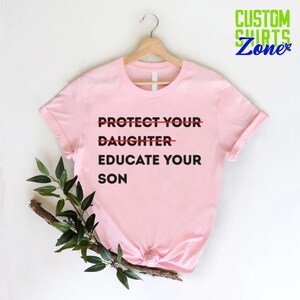 Pu&ograve; includere: Maglietta rosa con il testo "PROTECT YOUR DAUGHTER EDUCATE YOUR SON" con una linea rossa che attraversa la parola "DAUGHTER".