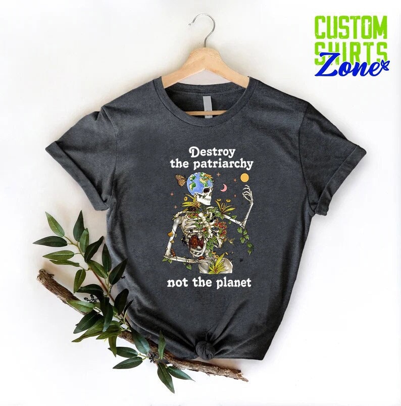 May include: Dark gray t-shirt with a skeleton holding the Earth, surrounded by vines and flowers. The text "Destroy the patriarchy not the planet" is printed on the shirt.