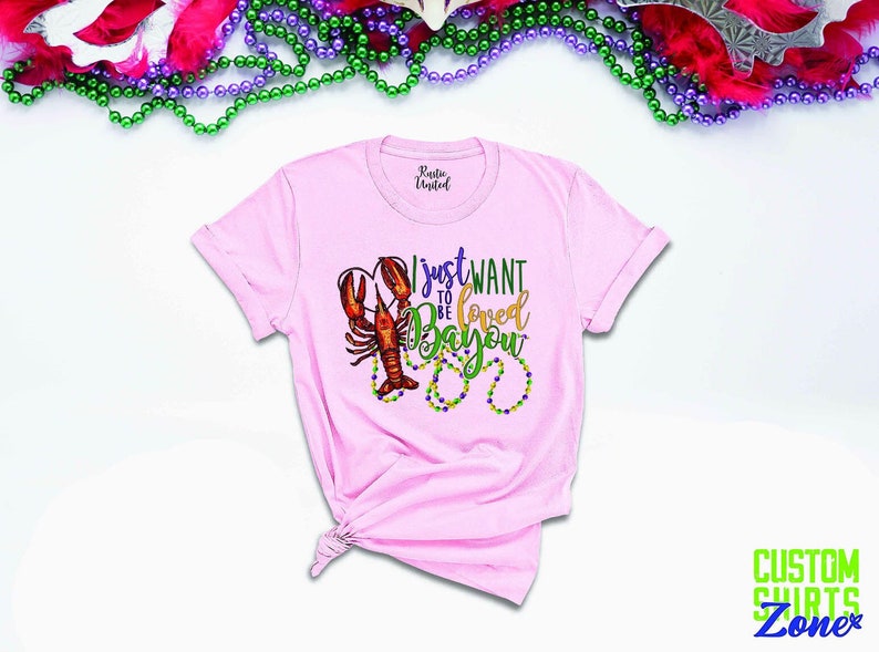 May include: Pink t-shirt with a graphic of a crawfish and Mardi Gras beads. The text on the shirt reads "I just want to be loved Bayou".