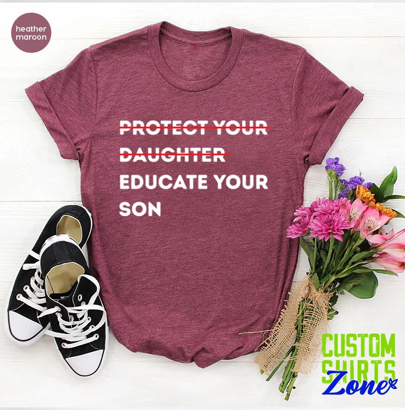Pu&ograve; includere: Una t-shirt color marrone rossiccio con la scritta "PROTECT YOUR DAUGHTER EDUCATE YOUR SON" stampata in bianco. La parola "DAUGHTER" &egrave; barrata. La t-shirt &egrave; appoggiata su una superficie bianca con un mazzo di fiori e un paio di scarpe da ginnastica nere.