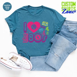 May include: A teal blue t-shirt with a pink heart and the text "I love the 80s" in a retro font. The heart has a disco ball inside and there are other 80s icons around the text, including roller skates, a cassette tape, sunglasses, and a boombox.