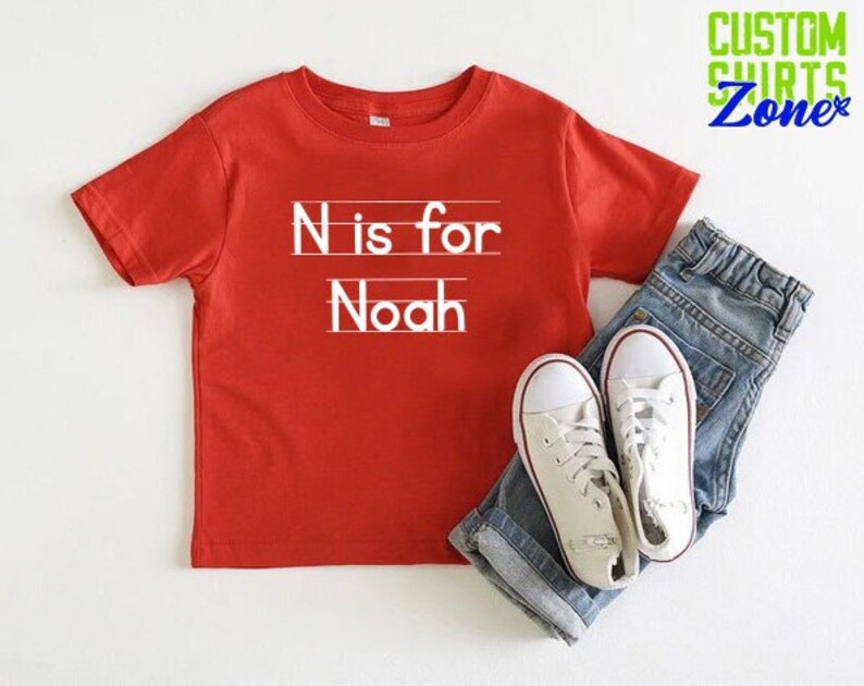 May include: Red short-sleeved t-shirt with white text that says "N is for Noah" on a white lined background.