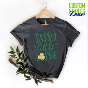 K&ouml;nnte beinhalten: Ein dunkelgraues T-Shirt mit dem Text "SASSY little LASSIE" in gr&uuml;ner Schrift. Neben dem Text befindet sich ein goldener Glitzer-Kleeblatt.