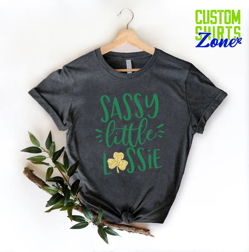 K&ouml;nnte beinhalten: Ein dunkelgraues T-Shirt mit dem Text "SASSY little LASSIE" in gr&uuml;ner Schrift. Neben dem Text befindet sich ein goldener Glitzer-Kleeblatt.