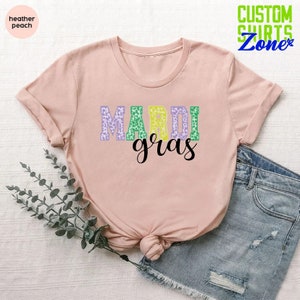 May include: A pink t-shirt with the words "Mardi Gras" in purple, green, and yellow leopard print letters. The shirt is laid flat on a white surface with a sprig of greenery and a pair of blue denim shorts.