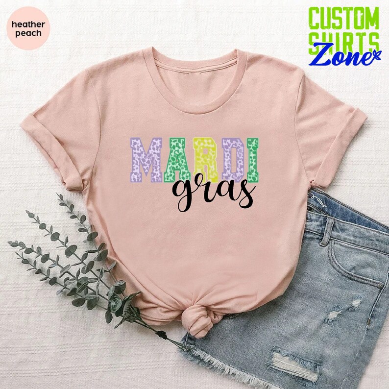 May include: A pink t-shirt with the words "Mardi Gras" in purple, green, and yellow leopard print letters. The shirt is laid flat on a white surface with a sprig of greenery and a pair of blue denim shorts.