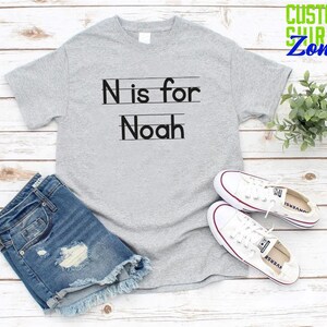 May include: Gray t-shirt with black text that says "N is for Noah" with lines underneath the letters. The shirt is on a white wood background with a pair of white sneakers and blue denim shorts.