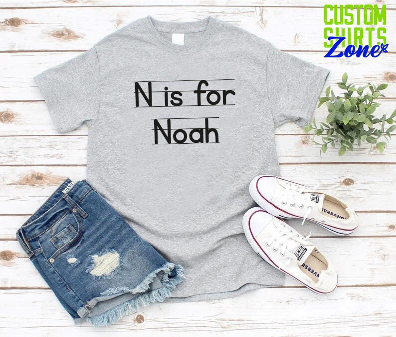 May include: Gray t-shirt with black text that says "N is for Noah" with lines underneath the letters. The shirt is on a white wood background with a pair of white sneakers and blue denim shorts.